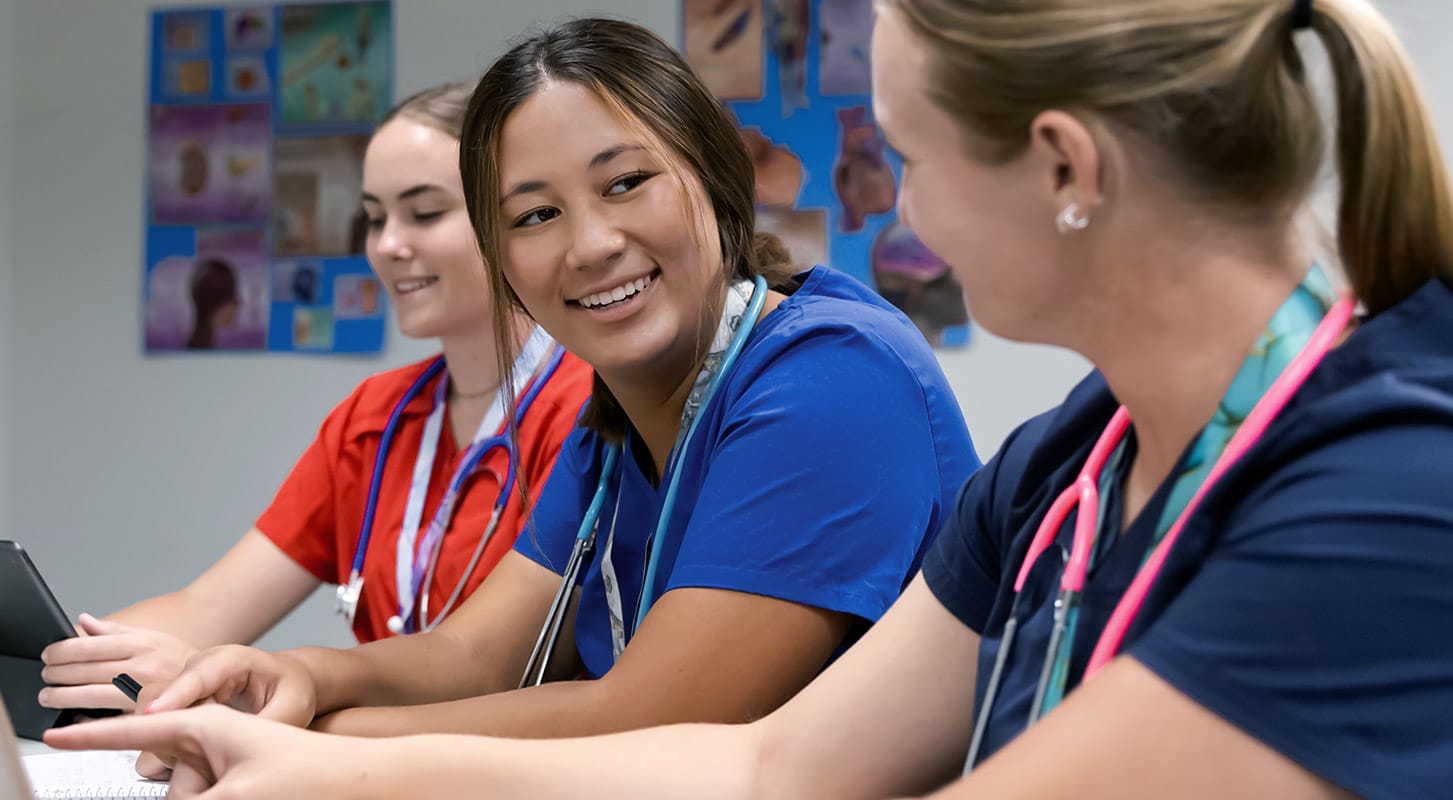 Three women in scrubs collaborate at a table, focused on a laptop in a professional setting.
